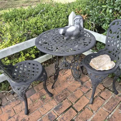 Three-piece cast metal bistro set featuring a round table with rose detail and two matching chairs, shown outdoors on a brick patio.