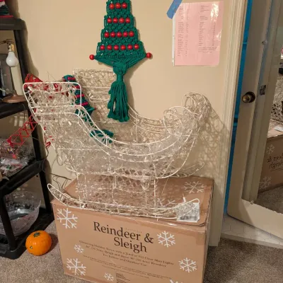 Lit wire Santa's sleigh Christmas decoration displayed on its original box, accompanied by a green macramé Christmas tree wall...