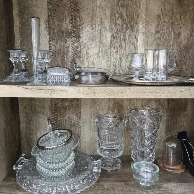 Collection of crystal and clear glassware displayed on wooden shelves, including cut glass vases, candlesticks, stemware, and serving...