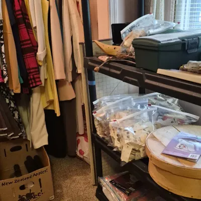 Black plastic shelving unit holding craft supplies, tackle boxes, and plastic bags alongside hanging vintage clothing.