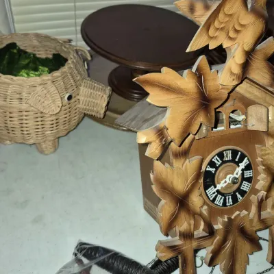 Hand-carved Black Forest style cuckoo clock featuring bird and leaf details, displayed alongside a pig-shaped wicker basket and a wooden...