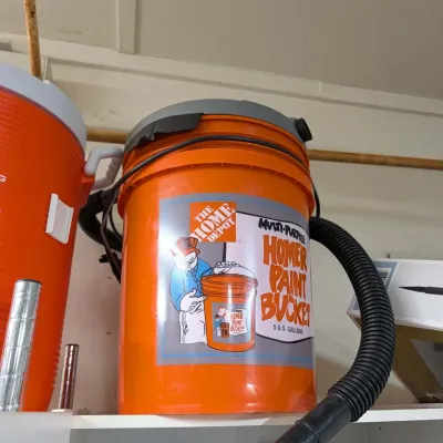 Home Depot Bucket Head wet/dry vacuum attached to a 5-gallon Homer paint bucket, shown sitting on a shelf next to an orange water cooler.