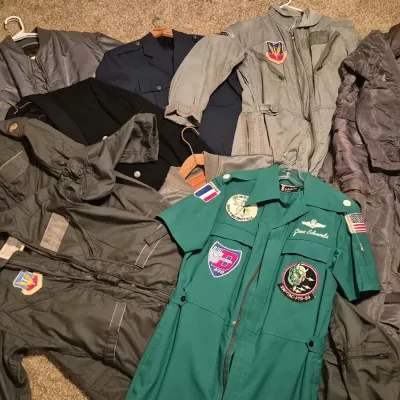 Collection of vintage military flight suits, dress uniforms, and jackets, including a teal short-sleeve suit with various unit patches.