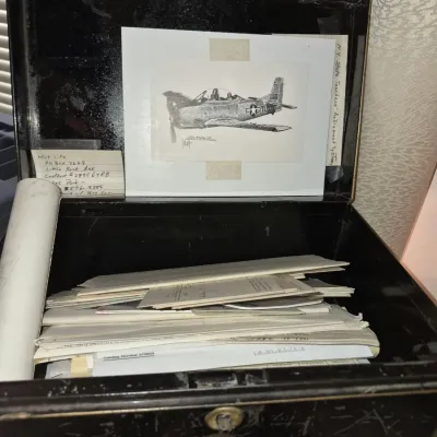 Vintage black metal lockbox containing personal documents, handwritten notes, and an airplane illustration taped inside the lid.
