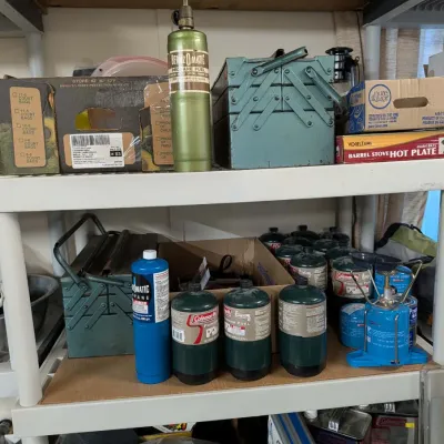 Workshop and camping supplies organized on plastic shelving, including a BernzOmatic propane torch, multiple propane canisters, green...