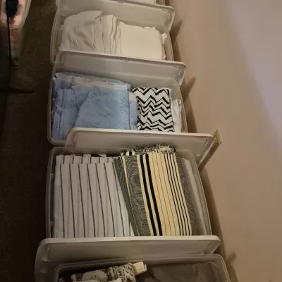 A row of clear plastic storage totes filled with folded fabrics, linens, and textiles including striped patterns and solid colors.