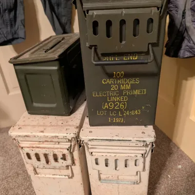 Stack of vintage military ammo cans, including a 1973 20mm olive drab metal ammunition container and painted surplus cans.