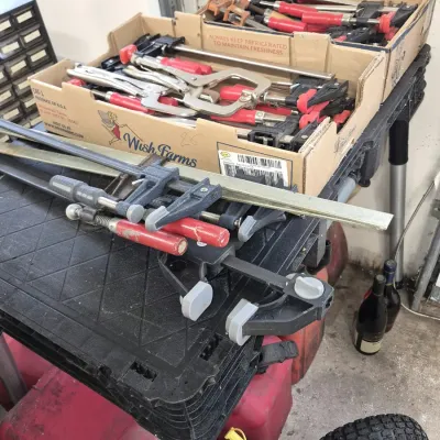 Large assortment of woodworking clamps, bar clamps, and locking pliers organized in cardboard flats atop a folding workbench.
