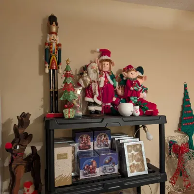 Collection of Christmas decorations displayed on shelving, including plush elves, a tall wooden nutcracker, boxed ornaments, and a wooden...