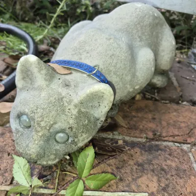 Weathered concrete garden statue of a crouching cat featuring glass insert eyes and a blue collar, resting on a brick patio.