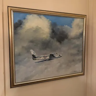 Framed oil painting depicting a military jet fighter soaring through clouds, housed in a silver and gold tone wooden frame.
