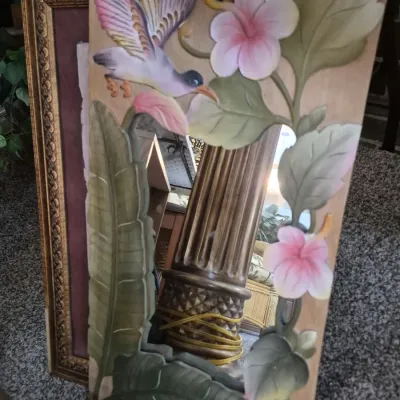 Hand-carved wooden mirror featuring painted tropical motifs of a bird and hibiscus flowers, leaning against a framed picture.