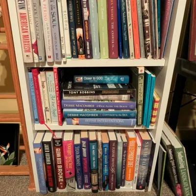 White wooden bookshelf filled with an assortment of hardcover and paperback books, featuring non-fiction, memoirs, and popular fiction...