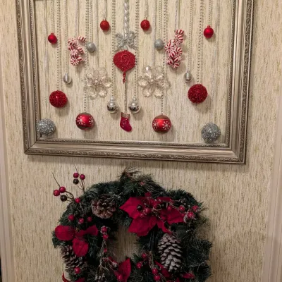 Framed Christmas ornament display with red and silver baubles suspended on beaded strings, mounted above a festive artificial poinsettia...