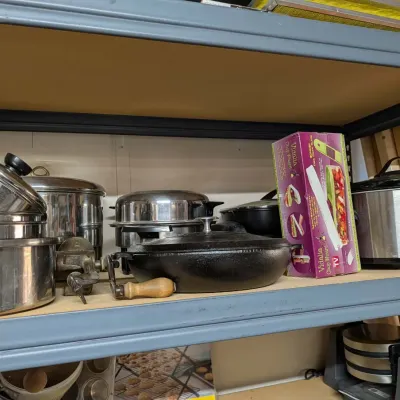 Assorted kitchenware arranged on a shelf, including stainless steel pots, a cast iron skillet with wooden handle, a hand-crank grinder, a...