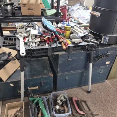 Assortment of workshop tools, including wrenches, clamps, and caulk guns, displayed alongside vintage metal storage trunks and shelving...