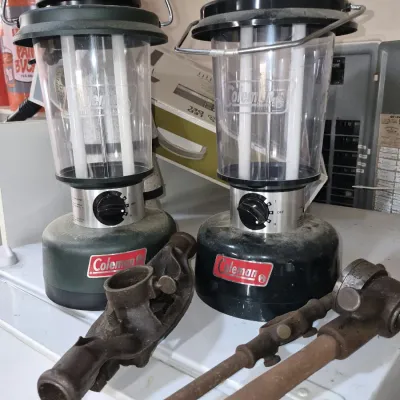 Pair of battery-operated Coleman fluorescent camping lanterns displayed alongside vintage rusted mechanical metal tool parts.