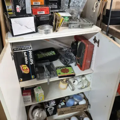 White storage cabinet filled with various household hardware and supplies, including light bulbs, screws, a bench vise, and electrical...
