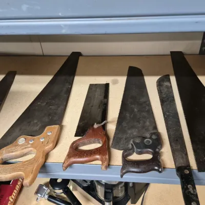 Collection of vintage hand saws and a machete, featuring carved wooden handles and aged steel blades arranged on a workshop shelf.