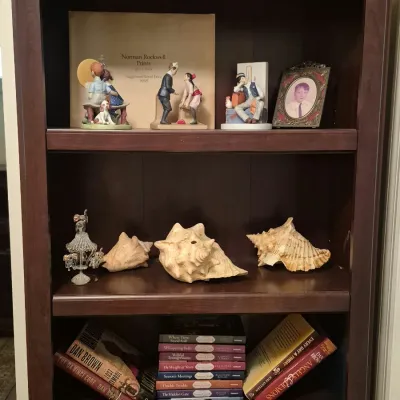 Dark wood bookcase filled with collectibles, including Norman Rockwell figurines, a framed portrait, large conch shells, and various...