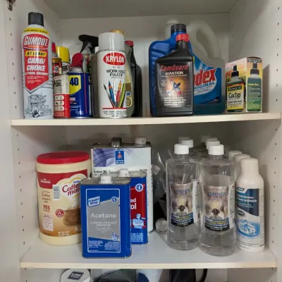 Assortment of household chemicals, automotive supplies, lamp oils, and shooting accessories arranged on white cabinet shelves.