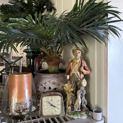 Sale Photo Thumbnail #241: Vintage ceramic figurine of a hunter with his dog, displayed alongside a small vintage alarm clock, candle in glass hurricane, and potted faux palm plant.