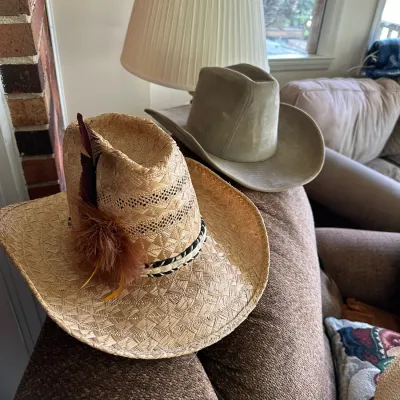 Sale Photo Thumbnail #630: Two cowboy hats resting on the back of a sofa, including a woven straw hat with a feather embellishment and a light gray felt cowboy hat.