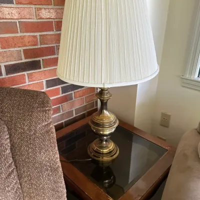 Sale Photo Thumbnail #641: Vintage brass table lamp with a pleated cream fabric shade, resting on a square wooden glass-top end table.