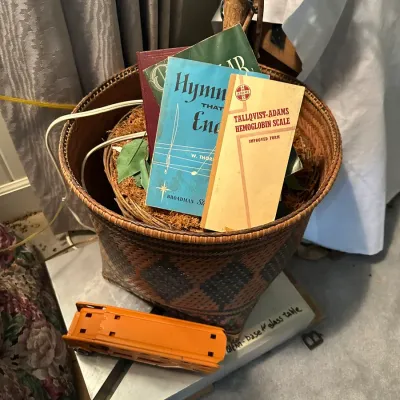 Sale Photo Thumbnail #368: Woven basket filled with vintage books and pamphlets, alongside a toy orange truck resting on a table below.