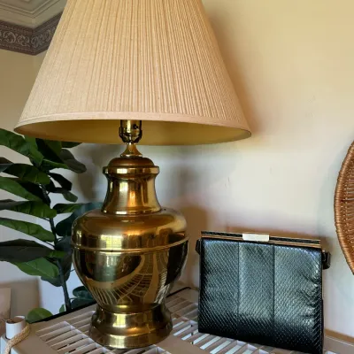 Sale Photo Thumbnail #677: Polished brass urn-style table lamp paired with a pleated beige fabric shade and a vintage black snakeskin clutch purse.