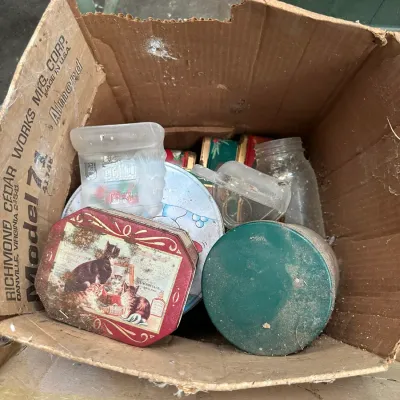 Sale Photo Thumbnail #561: Cardboard box containing vintage tins, clear glass jars, and plastic containers showing age and surface dust.