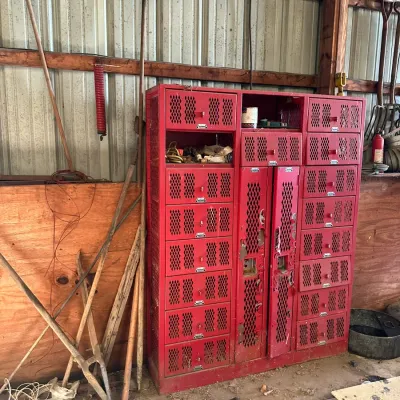 Sale Photo Thumbnail #567: Vintage red painted metal storage cabinet unit with perforated locker doors and small drawers, surrounded by assorted long-handled yard tools.