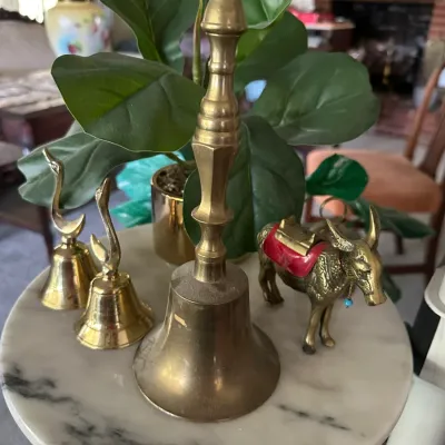 Sale Photo Thumbnail #243: Collection of vintage brass items on a marble table, including hand bells with decorative handles and a detailed ox figurine with red accents.