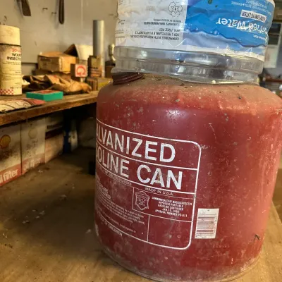 Sale Photo Thumbnail #480: Vintage red galvanized 5-gallon gasoline can with visible wear and surface distress, topped with a large plastic water jug.
