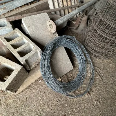 Sale Photo Thumbnail #332: Assortment of farm supplies including rolls of barbed wire, concrete cinder blocks, wire fencing, and metal pipes.