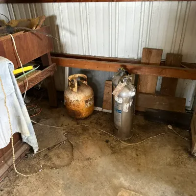 Sale Photo Thumbnail #470: A heavily rusted yellow propane tank alongside a stainless steel fire extinguisher stored on a concrete garage floor.