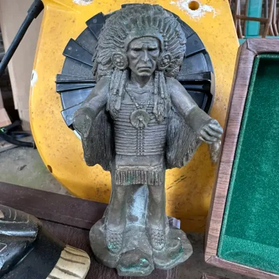 Sale Photo Thumbnail #158: Cast metal Native American chief figure with detailed feather headdress and breastplate, showing some aged patina and missing its right hand.
