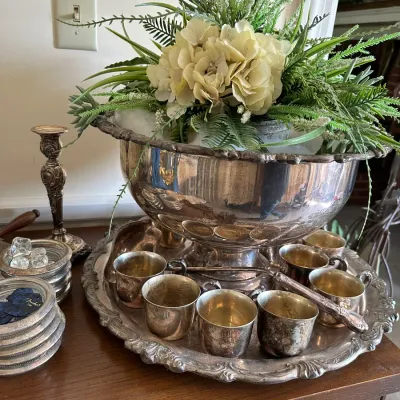 Sale Photo Thumbnail #668: Vintage silver-plated punch bowl set with matching tray, cups, ladle, and a decorative faux floral arrangement, displayed alongside additional silver accessories.