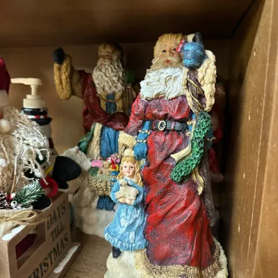 Sale Photo Thumbnail #305: Collection of resin Santa Claus figurines, including a tall Santa standing alongside a young girl in a blue dress holding a lamb, displayed on a wooden shelf.