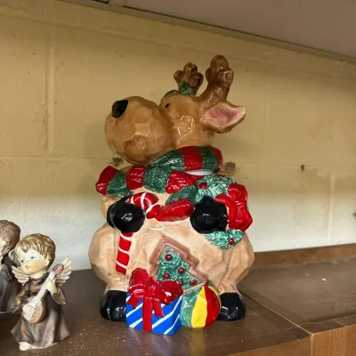 Sale Photo Thumbnail #306: Festive ceramic reindeer cookie jar featuring hand-painted holiday details including a scarf, wreath, candy cane, and presents, shown alongside small angel figurines on a wooden shelf.