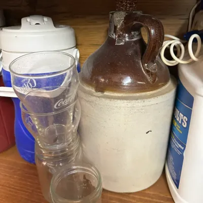 Sale Photo Thumbnail #88: Vintage two-tone stoneware whiskey jug with corncob stopper, displayed alongside vintage Coca-Cola glasses and clear glass jars on a shelf.