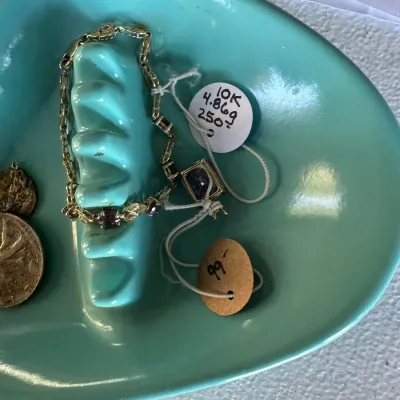 Sale Photo Thumbnail #409: A turquoise ceramic dish holding a 10K yellow gold link bracelet set with purple gemstones, paired with a matching pendant and two coins.