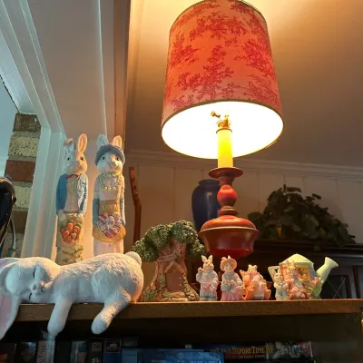 Sale Photo Thumbnail #195: Collection of decorative rabbit figurines and a table lamp with a red toile pattern shade, displayed atop a shelf alongside VHS tapes.