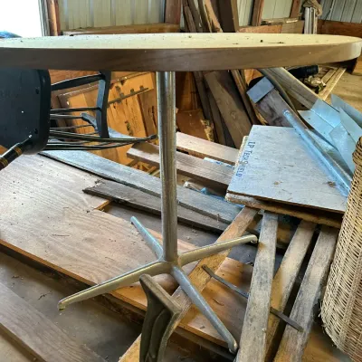 Sale Photo Thumbnail #539: Round mid-century bistro table featuring a wood or laminate top resting on a metal pedestal base, stored in a barn among miscellaneous materials.