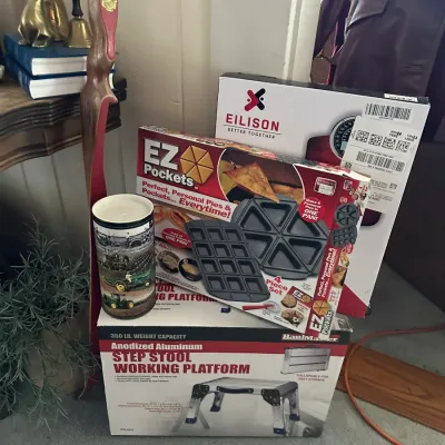 Sale Photo Thumbnail #249: Collection of household goods including a boxed HaulMaster step stool platform, EZ Pockets baking set, and Eilison box, alongside a vintage John Deere candle and brass decor.