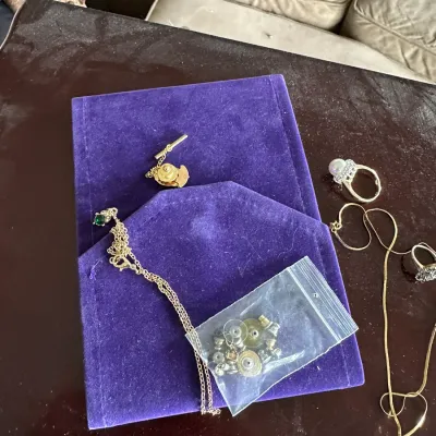 Sale Photo Thumbnail #138: Collection of fine and costume jewelry, featuring a pearl cocktail ring, gemstone pendant chains, tie bar, and a baggie of earring backs displayed on a purple velvet pouch.