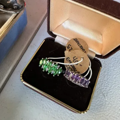 Sale Photo Thumbnail #407: Pair of gemstone cluster rings presented in a velvet jewelry box, including a marquise green stone cluster ring and a row-set purple amethyst band.