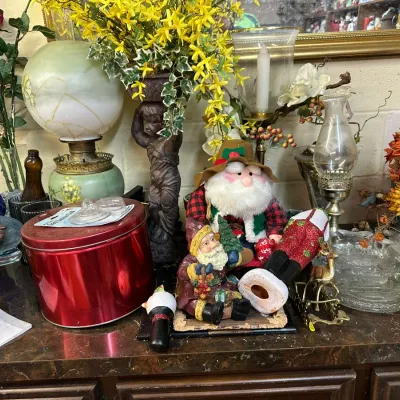Sale Photo Thumbnail #313: Collection of home decor including vintage oil lamps, holiday Santa figurines, floral arrangements, and a red metal tin displayed on a tabletop.