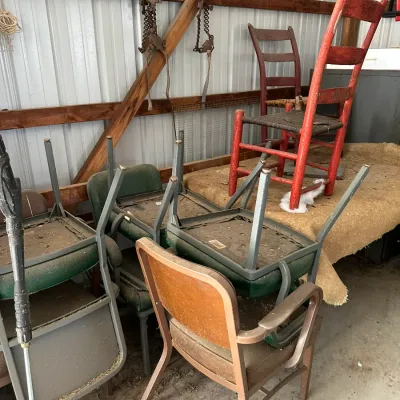 Sale Photo Thumbnail #718: Collection of vintage furniture in a barn, including mid-century metal-framed office chairs, a brown vinyl armchair, and rustic painted wooden ladder-back chairs.