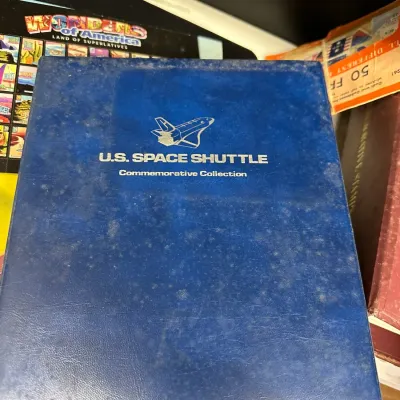 Sale Photo Thumbnail #93: Blue leatherette 'U.S. Space Shuttle Commemorative Collection' album binder displaying silver foil lettering and shuttle graphic, showing age-related surface wear.